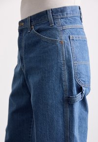 Dongeribukser i medium blå, med en rett ben-design, fem lommer, synlig søm, og en sideverktøylomme.