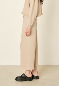Beige, cropped sweatshirt med lange ærmer, parret med løstsiddende, højtaljede beige bukser. Teksturen er blød og har rå kant detaljer. Sorte klipklapper.