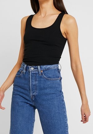 Femme portant un débardeur noir sans manches et un jean bleu taille haute sur un fond gris clair uni.