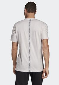 Lichtgrijze T-shirt met korte mouwen en een verticale zwarte tekstprint langs de achterkant. Gemaakt van een zachte, gladde stof.