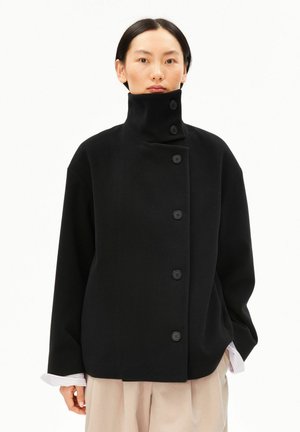 Personne portant un manteau noir à col montant boutonné avec des manches larges et un pantalon beige, debout devant un fond blanc uni.