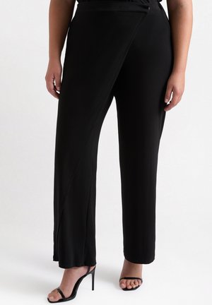 Schwarze weit geschnittene Hosen aus glattem Stoff, mit einem asymmetrischen Design an der Vorderseite und einem sauberen Saum. Kombiniert mit schwarzen High-Heels.