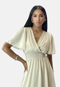 Beige dress with short flutter sleeves, v-neck, and smocked waist. Soft fabric, knee-length, with side pockets and a flowy silhouette.