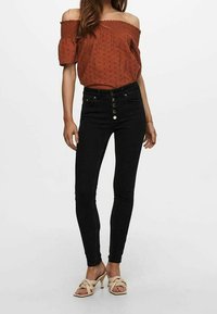Off-shoulder top i rustfarve med korte ærmer, parret med højtaljede sorte skinny jeans med frontknapper. Beige crisscross sandaler.