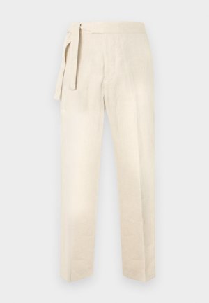 Άνδρας όρθιος με τα χέρια στις τσέπες, φοράει σκοτεινό γκρι πουκάμισο με γυρισμένα μανίκια, μπεζ παντελόνι με ζώνη και άσπρα αθλητικά παπούτσια.