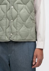 Πρασινός γούνινος γιλέκος με κυματοειδές σχέδιο, χαρακτηρισμένος από ασημένιους κουμπιά ύφους και δύο τσέπες στο στήθος. Φορεμένος πάνω από ένα ελαφρύ πουκάμισο με μακριά μανίκια και γκρίζα παντελόνια.