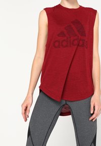 Punainen hihaton urheilu-toppi, jossa on iso musta "adidas"-logo, rento malli ja korkea-alemmainen helma, yhdistettynä harmaisiin tiukkoihin legginsseihin.