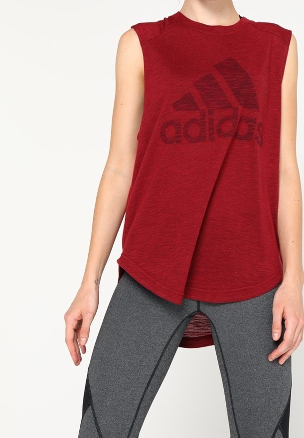 Punainen hihaton urheilu-toppi, jossa on iso musta "adidas"-logo, rento malli ja korkea-alemmainen helma, yhdistettynä harmaisiin tiukkoihin legginsseihin.