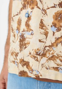 Camicia a maniche corte in tessuto beige chiaro con motivi di foglie marroni e blu, con un bottone blu e un orlo dritto.