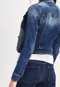 Jasno-niebieska kurtka dżinsowa o dopasowanym kroju, z delikatnym wyblakłym efektem, mosiężnymi elementami i zapięciem na guziki. Posiada klasyczny kołnierz oraz kieszenie na piersiach.