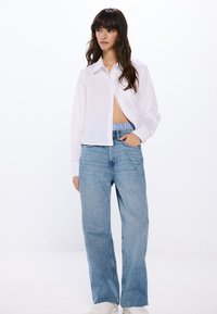 Camicia bianca a maniche lunghe con un design accorciato, abbinata a jeans in denim blu chiaro larghi. Tessuto liscio con una consistenza morbida. Calzature bianche.