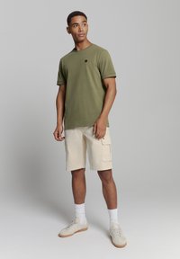 Olijfgroene katoenen t-shirt met korte mouwen, beige cargoshorts en beige sneakers. Model staat tegen een neutrale achtergrond.