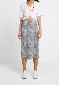 Weißes Kurzarm-Top mit gedrehtem Detail an der Taille, kombiniert mit einem schwarz-weißen gemusterten Midirock, abgerundet mit weißen Sneakern.