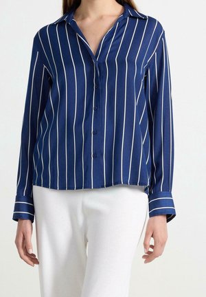 Kobieta w granatowej koszuli na guziki z białymi pionowymi paskami i białych spodniach, stojąca na jednolitym tle.