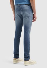 Lichtblauwe jeans met een vervaagd ontwerp, voorzien van een rechte pasvorm, achterzakken en een logopatch op de tailleband. Een informele schoeiselcombinatie is zichtbaar.