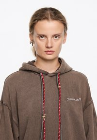 Sudadera con capucha marrón de corte holgado, cordones rojos y grises, y detalle de logo bordado. Textura suave y diseño casual.