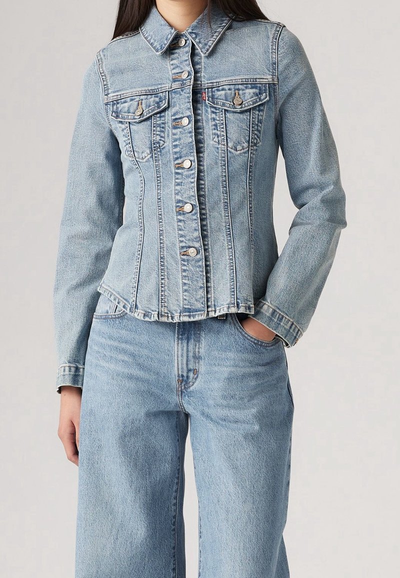 Femme portant une veste en denim bleu clair ajustée avec fermeture à boutons et jean taille haute assorti, main dans la poche, sur fond neutre.