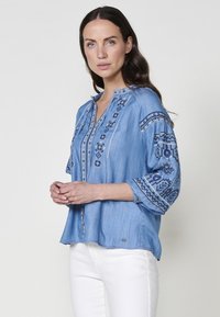 Ljusblå denimskjorta med broderade mönster i mörkare blått, med en henleykrage och trekvartslånga ärmar, avslappnad passform.