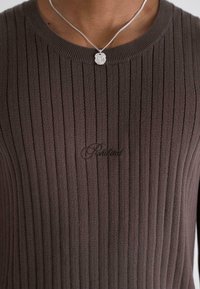 Brown ribbed sweater with a round neckline, featuring subtle logo embroidery on the front and a smooth texture. Silver necklace accent included.