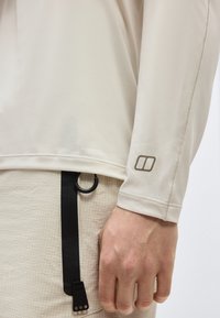 Κοντινό πλάνο του χεριού ενός ατόμου που φοράει μπεζ πουκάμισο με μακριά μανίκια και ένα μικρό λογότυπο κοντά στον καρπό, καθώς και υφασμένα ανοιχτόχρωμα παντελόνια με μαύρο λουράκι.