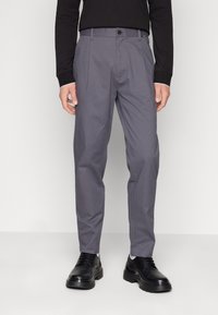 Pantaloni grigi a gamba dritta, con pieghe frontali. Abbinati a scarpe nere, che mostrano un tessuto liscio e un design sartoriale.