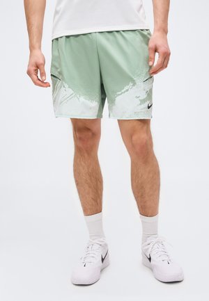 Férfi világoszöld és fehér mintás sportnadrágban, fehér zokniban és fehér Nike tornacipőben, egyszínű háttér előtt áll.