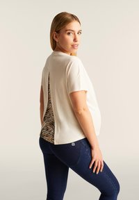 Top a maniche corte color crema con schiena aperta che rivela una stampa animale in marrone e nero. Abbinato a jeans blu scuro, che mostrano una vestibilità aderente.