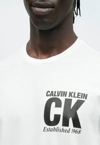 Biały bawełniany t-shirt z dużym czarnym logo "CK" i napisem "CALVIN KLEIN Established 1968" w pogrubionej czcionce. Prosty design z okrągłym dekoltem.