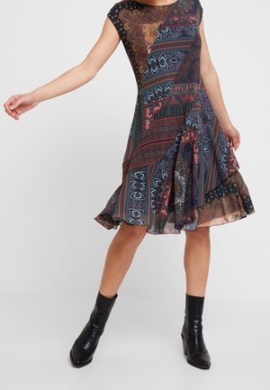 Femme portant une robe sans manches mi-longue à motifs géométriques multicolores et des bottines noires, debout les bras écartés.