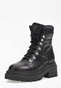 Bota de combate de cuero negro con una suela gruesa tipo plataforma, cordones en la parte delantera, herrajes plateados y un cuello acolchado. Cuenta con un parche con logo en el lateral.