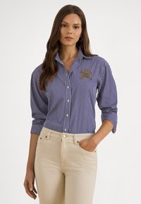 Sieviete ar gariem brūniem matiem, valkājot zilu un baltu svītrainu pogājamu kreklu ar emblēmu un bēšas krāsas bikses, viena roka kabatā.