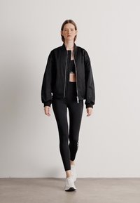 Fekete bomber dzseki egy rövid fekete top felett, fekete leggings-szel és fehér sportcipővel, egy semleges háttér előtt állva.