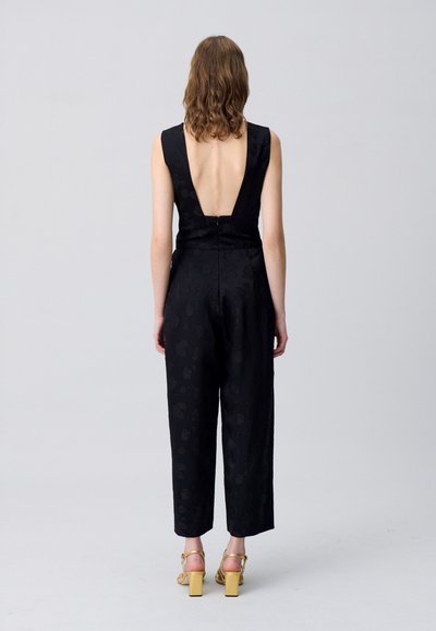 Jumpsuit nero senza maniche con scollo a V profondo, motivo floreale testurizzato e gambe affusolate. Indossato con sandali con tacco a blocco dorati.