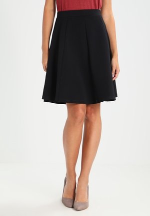 Femme portant une jupe noire évasée mi-longue, un haut rouge et des chaussures à talons beiges, debout sur un fond uni.