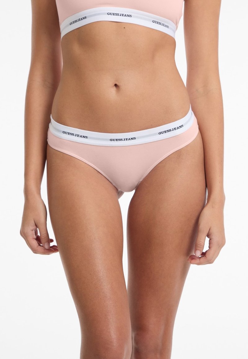 Lichtroze bikini met een laag uitgesneden pasvorm, met een witte elastische tailleband waarop "GUESS JEANS" in zwart gedrukt staat. Gladde textuur, strakke pasvorm.
