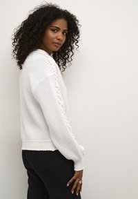 Maglione bianco lavorato a maglia con orlo a coste e dettagli di volant lungo le maniche, abbinato a pantaloni neri. Tessuto liscio, vestibilità casual.