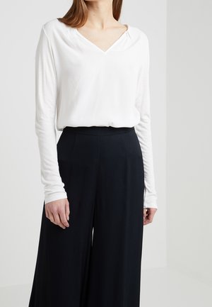 Kvinde iført hvid top med lange ærmer og V-hals, stoppet ned i sorte bukser med højtaljede, brede ben mod en ensfarvet baggrund.