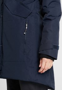 Gros plan sur la manche d'un manteau d'hiver bleu marine avec une poche zippée, une main partiellement visible à côté du poignet.
