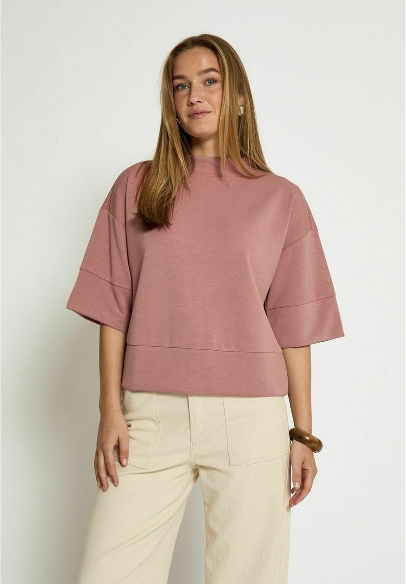 Top rosa a maniche corte con scollatura rotonda, design corto e forma quadrata. Il tessuto ha una consistenza liscia, abbinato a pantaloni larghi di colore chiaro.