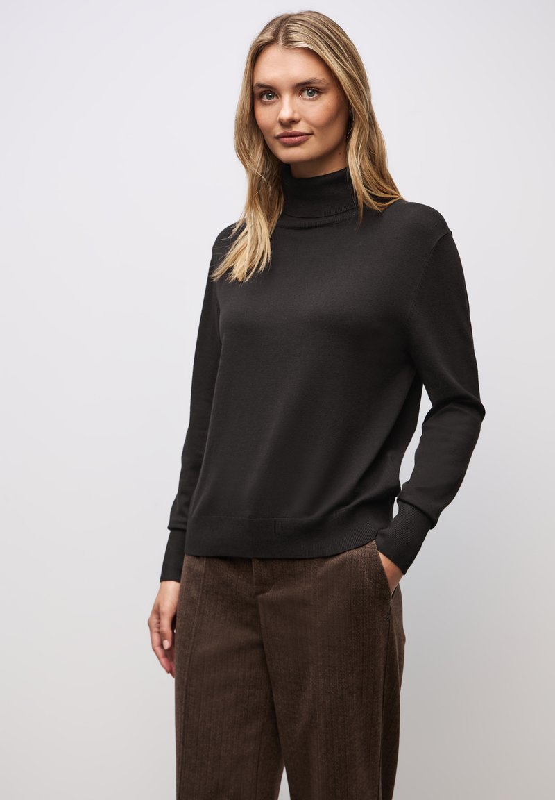 Schwarzer Rollkragenpullover aus weichem Strickmaterial, lässige Passform, gerippte Bündchen und Saum, kombiniert mit braunen strukturierten Hosen, das Modell hat die Hände in den Taschen.