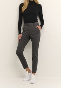 Pantaloni grigi su misura con tasche laterali, taglio dritto e una consistenza liscia, abbinati a un top nero a maniche lunghe e sneaker bianche.