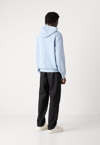 Sudadera azul claro con capucha, combinada con pantalones oscuros y holgados y zapatillas beige, vista desde la espalda sobre un fondo liso.