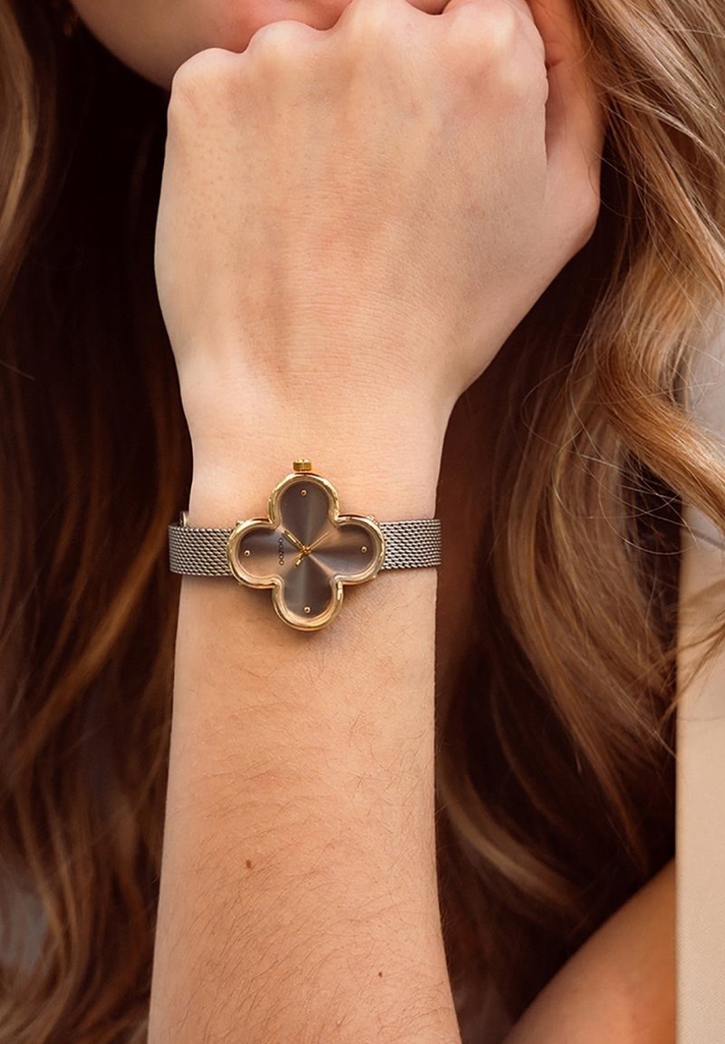 Montre-bracelet en métal doré avec un cadran en forme de trèfle, un cadran gris, un bracelet en mailles et quatre petits marqueurs d'heure.