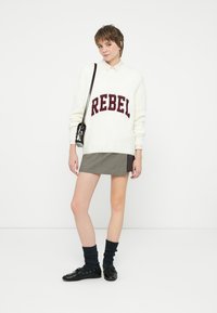 Bílý svetr s nápisem "REBEL" v burgundské barvě, doplněný kostkovanou mini sukní. Černé boty a tmavé strukturované ponožky dotvářejí vzhled.