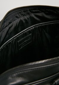 Wnętrze czarnej skórzanej torby z zamkiem błyskawicznym, gładką podszewką z tłoczonym logo oraz wewnętrzną kieszenią z przyszytą metką.