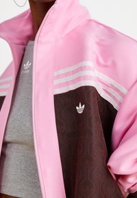 Rosa idrottsjacka med brun nederdel, som har tre vita ränder på axlarna och logotypbroderi på bröstet.