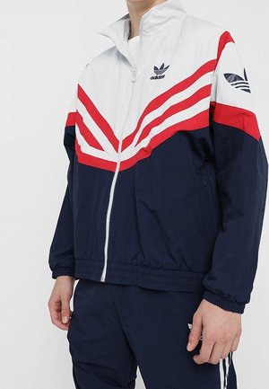 Mand iført en marineblå, hvid og rød Adidas træningsjakke med fuld lynlås, logo på bryst og ærme, kombineret med marineblå bukser.