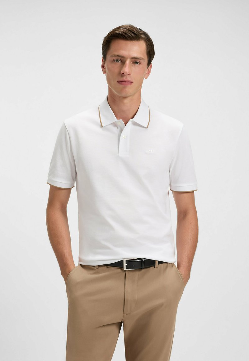 Vit polo skjorta med slim fit, med en kontrastrande brun krage och manschetter, tillverkad av mjukt tyg. Buren med beige byxor och ett svart bälte.