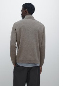 Pull en maille texturée marron avec un col haut, des manches longues et une coupe décontractée, associé à une chemise bleu clair en dessous.