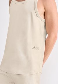 Débardeur beige en coton doux avec un col rond et une bordure cousue. Présente un petit logo et un détail textuel sur l'ourlet.